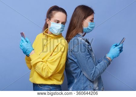 Picture Of Two Busy Curious Attractive Females Standing Back To Back Holding Their Smartphones, Usin