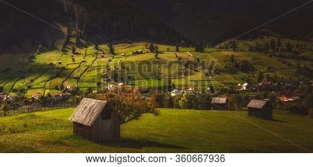 Panorama View Of A Mountain Village At Sunset. Household With Meadows And Farmland