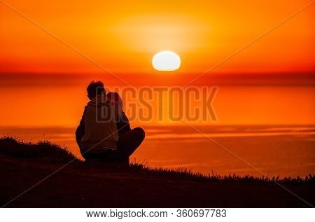 A Red Burning Sunset Over The Sea With The Silhouette Of A Young Couple Enjoying Evening