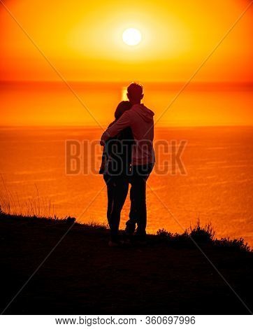 A Red Burning Sunset Over The Sea With The Silhouette Of A Young Couple Enjoying Evening