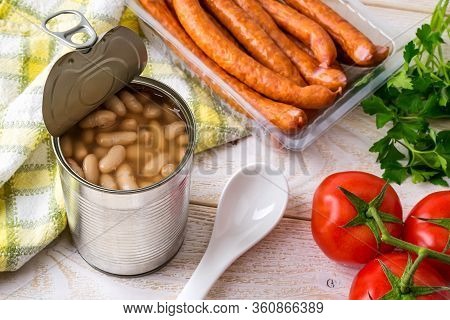 Canned Backed Beans In An Open Tin Can With Pull Tab Near Thin Sausages, Tomatoes And Ceramic Spoon 