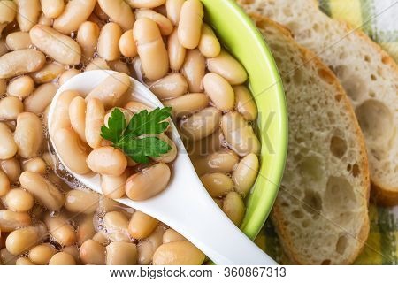 Close-up Of White Backed Beans In A Ceramic Spoon In A Green Bowl Near Slices Of Bread. Bean Is Sour