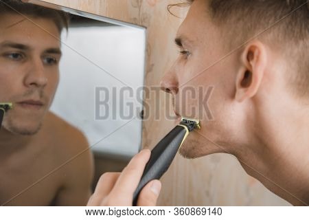Close Up Of Young Man Looking To Mirror And Shaving Beard With Trimmer Or Electric Shaver