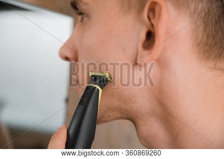 Close Up Of Young Man Looking To Mirror And Shaving Beard With Trimmer Or Electric Shaver