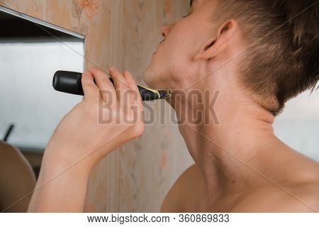 Close Up Of Young Man Looking To Mirror And Shaving Beard With Trimmer Or Electric Shaver