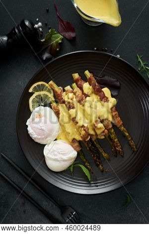 Baked Aspargus Covered With Bacon And Poached Eggs On A Black Ceramic Plate