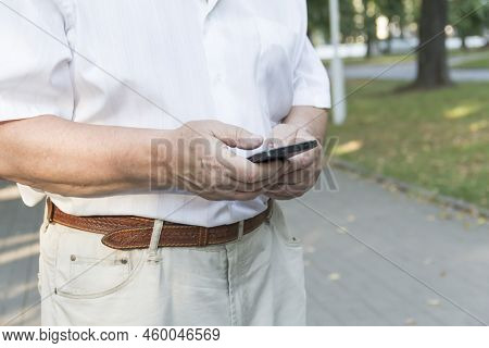 A Mobile Phone In The Large Hands Of An Elderly Man In A White Shirt On A Walk In The Park.