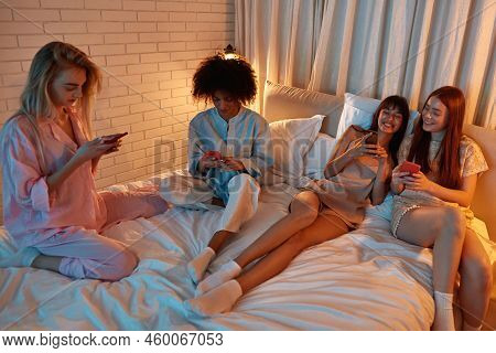 Young Multiethnic Girlfriends Using And Watching Smartphones On Bed During Girlish Sleepover. Black 