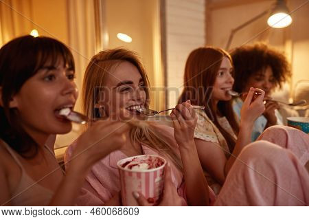 Young Multiracial Girlfriends Eat Ice Cream And Watch Tv Or Movie On Sofa During Girlish Sleepover A