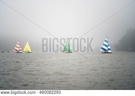 Four Colorful Sailboats Lost In The Haze Fog With Smaller Pedal Row Boats Floating Around On The Clo