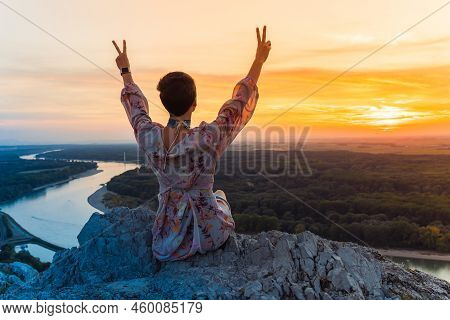 Young Teenager Girl Sits On Edge Of Stone Cliff, View From Back, Contemplates Beautiful Landscape, L