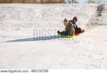 A Boy And A Girl Ride Down A Hill On A Plastic Sled. The Children Go Down The Hill On The Ice. Winte