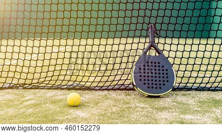 Professional Padel Racket And Yellow Ball Behind Net On Green Court Grass Turf
