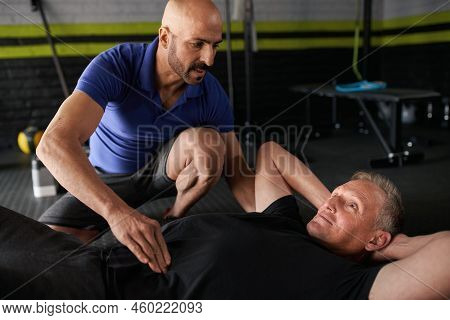 Male Personal Trainer Assisting An Elderly Mature Man In Doing Ab Crunches. Working On Core Strength