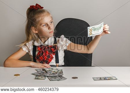A Little Girl Sits At A Table In Front Of A Stack Of Hundred-dollar Bills And Examines One Of Them F