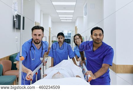 Doctors And Nurses Wheeling Elderly Patient In Gurney On Hospital Corridor At Emergency Department. 