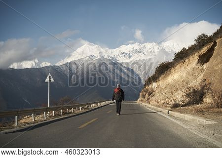 Rear View Of An Asian Hiker With Backpack Walking On Highway With Snowcapped Meili Mountain In Backg