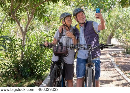 Active Caucasian Senior Couple With Electrobikes Standing Outdoors In The Park Video Calling By Cell