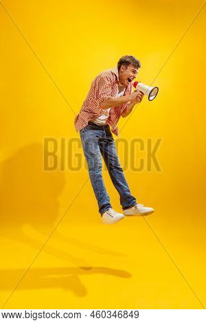 Portrait Of Young Man In Checkered Shirt Posing, Shouting In Megaphone In A Jump Isolated Over Yello