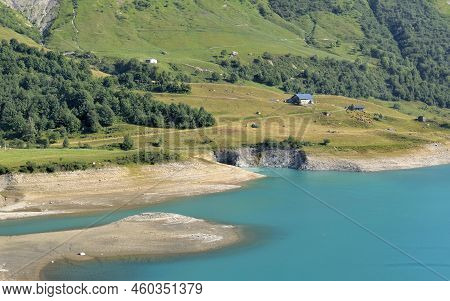 Riverbank Of The Lake Of Roselend And Blue Turquoise Water With A Farm Far Away At The Foot Of The M