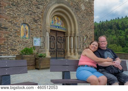 Smiling Middle-aged Tourist Couple Sitting And Hugging On Bench Against Stone Wall And Entrance Of C
