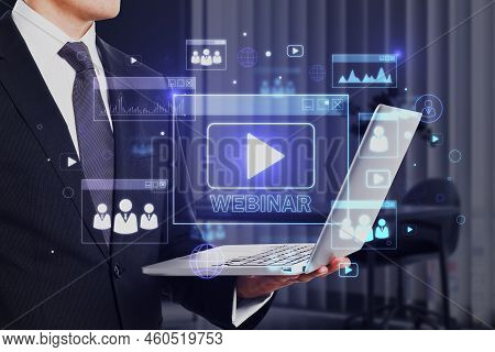 Close Up Of Businessman Hand Holding Laptop With Glowing Webinar Hologram On Blurry Office Interior 