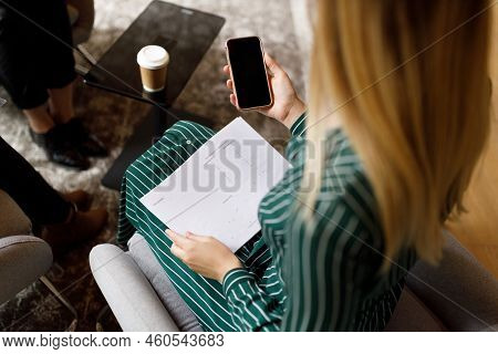 High Angle View Of Young Businesswoman With Blond Hair Holding Report And Using Smartphone While Sit