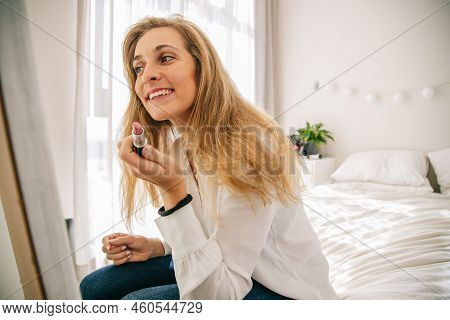 Beautiful Mid Adult Caucasian Woman With Blond Hair Holding Lipstick And Smiling While Looking In Mi