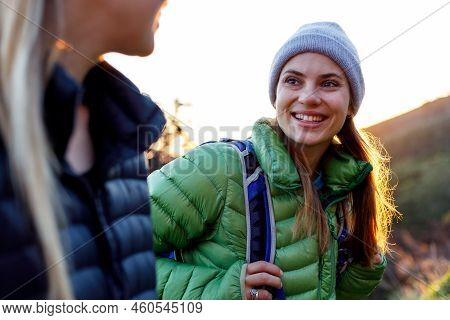 Smiling Young Woman In Down Jacket Talking With Female Friend While Hiking Against Clear Sky During 