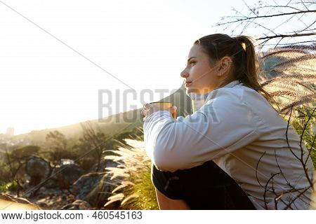 Thoughtful Young Woman Holding Coffee Cup And Looking Away While Relaxing On Mountain Against Clear 