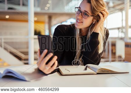 Young Blonde Caucasian Female Student Making A Video Call With Her Mobile Phone And Earphone To Her 