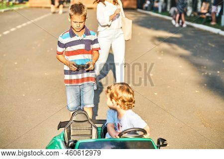 Elder Brother Controls Children Car With Remote At Walk With Little Brother In Public Park. Toddler 
