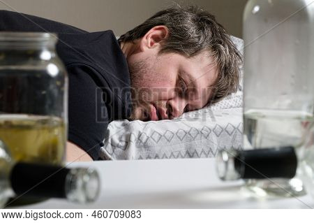 A Drunken Alcoholic Man Lies On The Bed, Empty Bottles On The Table. Alcoholism, Alcohol Dependence 