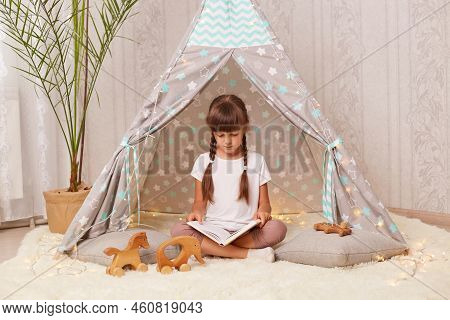 Full Length Portrait Of Little Girl With Braids In White T Shirt Reading Interesting Book While Sitt