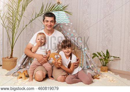 Portrait Of Optimistic Cheerful Family Wearing White T Shirts Sitting In Wigwam, Looking Smiling At 