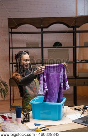 Smiling African Americn Designer Holding T-shirt Near Container In Workshop.