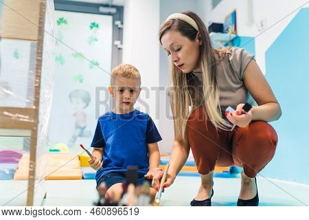 Nursery School. Caucasian Toddler Boy And His Teacher Painting On A Cling Film Wrapped Around The Wo