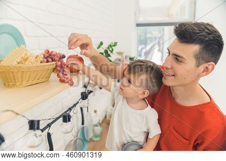 Happy Smiling Caucasian Brunet Dad Taking Snacks From The Top Kitchen Shelf With His Son. Grapes. Ap