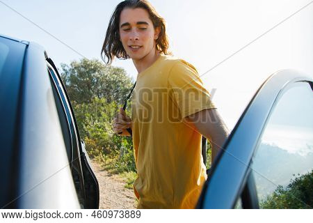 Young Male Traveler With Long Hair Standing By Car While Enjoying Vacation During Sunny Day
