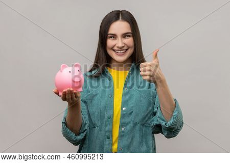 Portrait Of Smiling Pleased Woman With Dark Hair Holding Pig Money Box In Hands, Saving, Showing Thu