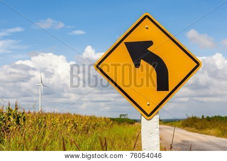 Left Curved Warning Sign With Windmill Background In Wind Farm, Thailand.