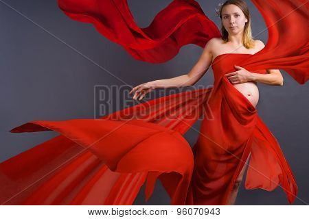 Portrait Of A Young Pregnant Woman In Red Dress With Blowing On Wind Waving Fabric, Grey Background,