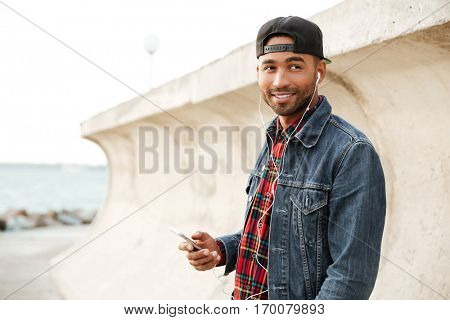 Happy african man wearing cap walking on the beach and chatting by his phone while listening music with earphones.