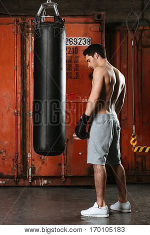 Image of handsome young strong boxer training in a gym. Looking aside. Standing near punchbag.