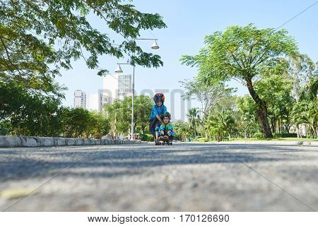 Cute little boy in helmet and knee pads riding skateboard while sitting on it, his elder brother pushing him down the road, surface level shot
