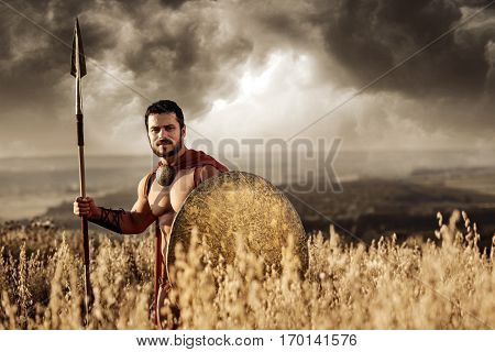 Warrior man with bare torso wearing red cloak holding sword and standing among field at dark. Male wearing like spartan or antique roman soldier going on war. Looking at camera.