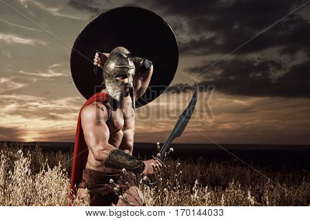 Warrior wearing in red cloak like spartan or antique roman soldier going forward in attack with iron sword. Incognito in helmet with bare torso holding bronze sword over head. Dark sky at sunset.
