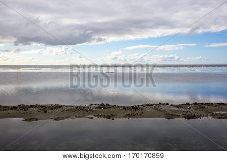 shoreline of Ameland Island with view over the wadden sea with clouds and tourmented sky reflecting in water at dawn