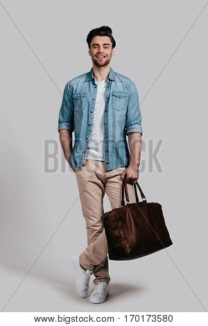 Stylish handsome man. Full length of good looking young man in casual wear carrying brown leather bag and smiling while standing against grey background
