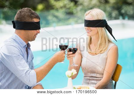 Blindfolded couple toasting red wine while sitting at poolside